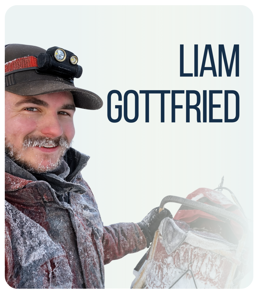 Person in cold weather gear with snow on face, holding a backpack, wearing a headlamp.