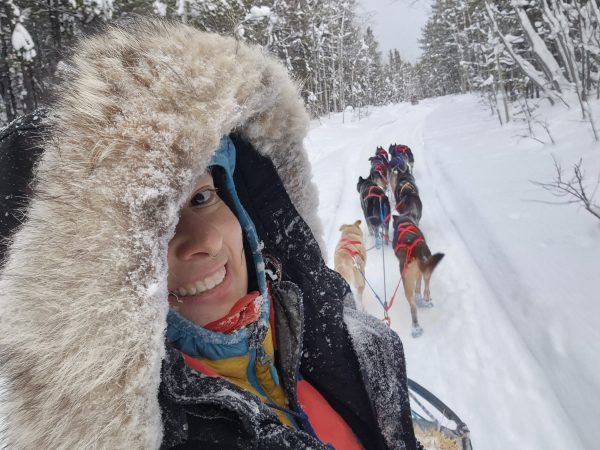 Person in fur-trimmed hood on snowy landscape with sled dogs in the background.