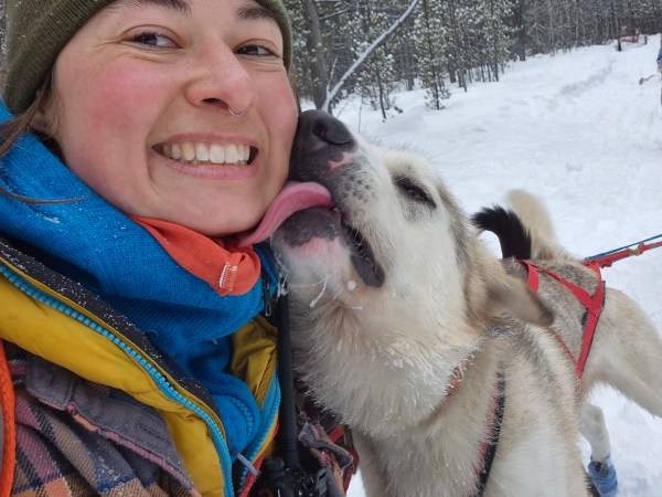 Person smiling as dog licks their face in snowy forest setting.