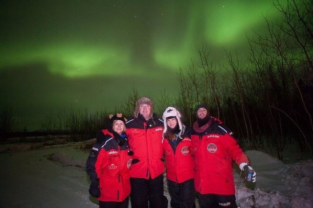 People in red jackets under the green glow of the Northern Lights.