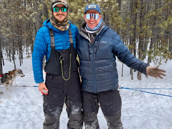 Two people in winter gear stand on snow, smiling, with trees in the background.
