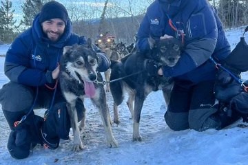 Two people in winter gear kneel with sled dogs in a snowy landscape.