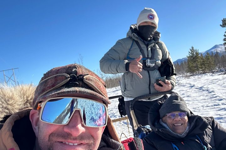 Three people wearing winter clothes pose outdoors on a snowy day with a clear blue sky.