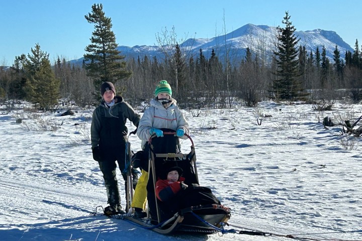 Two people pulling a sled with a child in snowy landscape, mountains in background.