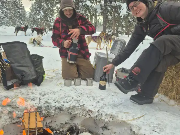 Two people in snowy forest setting by a campfire, preparing drinks with sled dogs in the background.